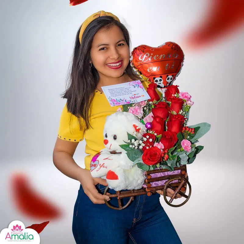 Joven sonriente sujetando un arreglo floral romántico en una carreta miniatura, con rosas rojas, un osito de peluche y un globo en forma de corazón.