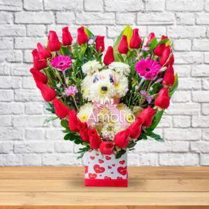 Arreglo floral con cachorro de flores blancas rodeado de rosas rojas y flores púrpuras, presentado en una base con corazones, ideal para expresar amor y ternura.