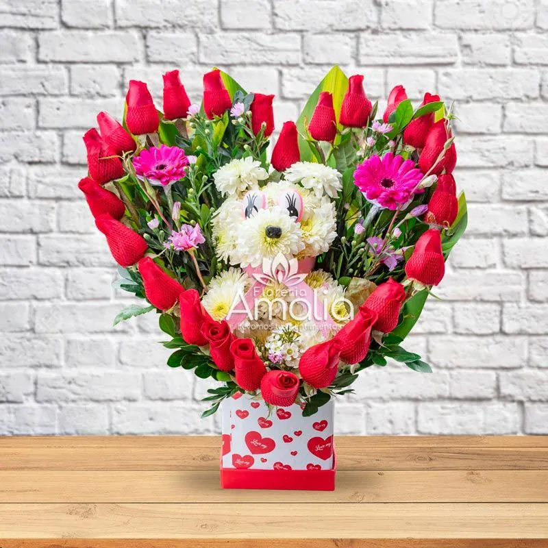 Arreglo floral con cachorro de flores blancas rodeado de rosas rojas y flores púrpuras, presentado en una base con corazones, ideal para expresar amor y ternura.