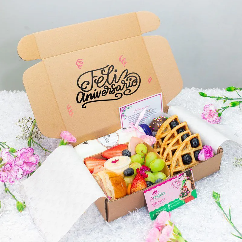 Caja de desayuno para aniversario con mensaje ‘Feliz Aniversario’, incluye frutas frescas, waffles, chocolates decorados, jugo natural, pan artesanal y tarjeta personalizada.