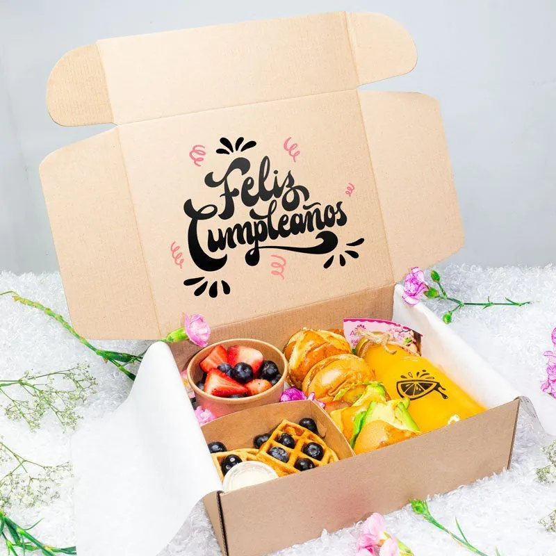 Caja de desayuno sorpresa con mensaje ‘Feliz Cumpleaños’, incluye frutas frescas, waffles, jugo natural, sándwiches y decoración floral.