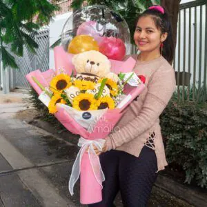 Ramo de girasoles con peluche, globos personalizados y decoración rosa, sostenido por una persona sonriente, ideal para regalos de cumpleaños o aniversarios