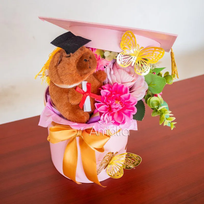 Caja decorativa de graduación con peluche, flores en tonos rosados y mariposas doradas.