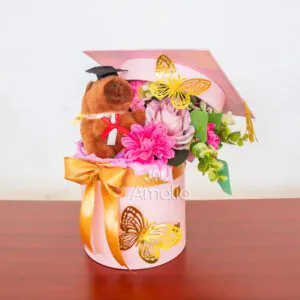 Caja decorativa de graduación con peluche, flores en tonos rosados y mariposas doradas.