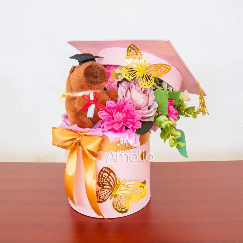 Caja decorativa de graduación con peluche, flores en tonos rosados y mariposas doradas.