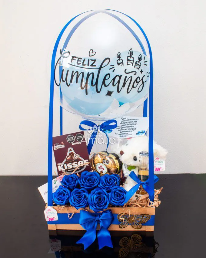 Caja de cumpleaños con globo personalizado, rosas azules, peluche blanco y dulces variados.