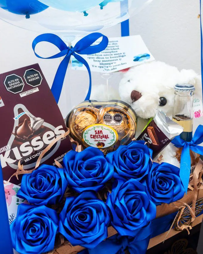 Caja de cumpleaños con globo personalizado, rosas azules, peluche blanco y dulces variados.