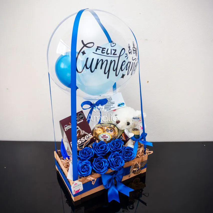 Caja de cumpleaños con globo personalizado, rosas azules, peluche blanco y dulces variados.