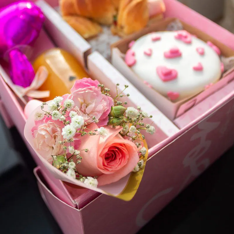 Desayuno sorpresa en caja rosa con mini ramo de flores, croissant relleno, jugo natural y torta decorada con corazones, ideal para regalos especiales.