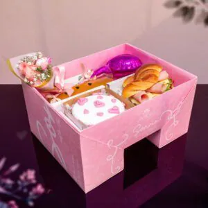 Desayuno sorpresa en caja rosa con mini ramo de flores, croissant relleno, jugo natural y torta decorada con corazones, ideal para regalos especiales.