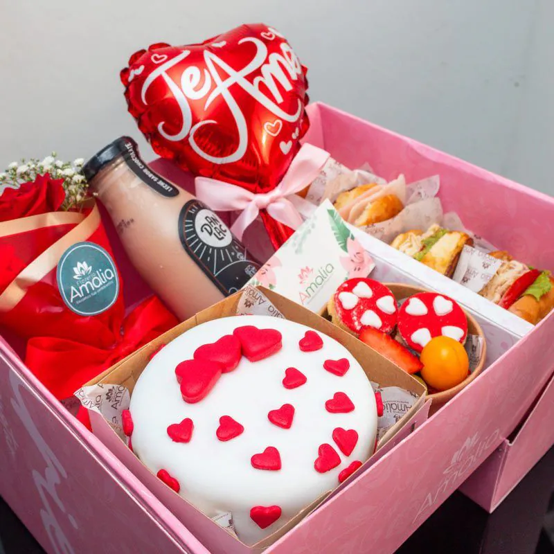 Desayuno sorpresa en caja rosa con mini torta decorada con corazones, sándwiches, jugo, flores y globo temático de ‘Te Amo’.