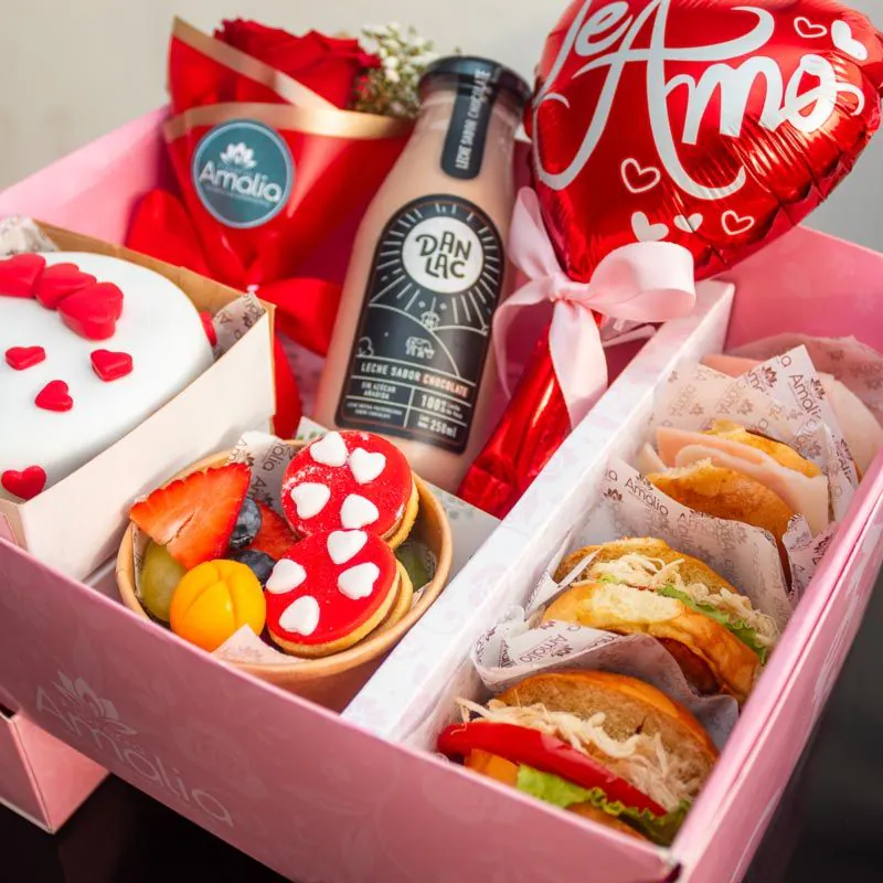 Desayuno sorpresa en caja rosa con mini torta decorada con corazones, sándwiches, jugo, flores y globo temático de ‘Te Amo’.