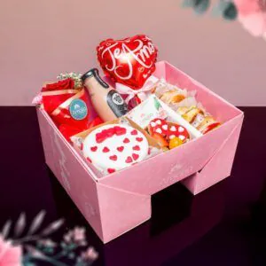 Desayuno sorpresa en caja rosa con mini torta decorada con corazones, sándwiches, jugo, flores y globo temático de ‘Te Amo’.