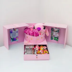 Caja sorpresa personalizada con rosas fucsias, peluche rosa tipo conejita, chocolates Hershey’s y dulces variados, ideal para sorprender con amor.