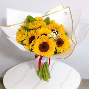 Ramo de girasoles frescos envuelto en papel blanco con borde dorado y decorado con mariposa dorada, ideal para regalar alegría y energía positiva.