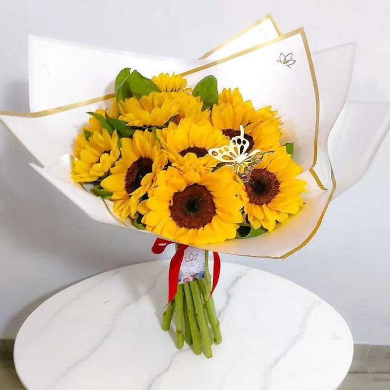 Ramo de girasoles frescos envuelto en papel blanco con borde dorado y decorado con mariposa dorada, ideal para regalar alegría y energía positiva.