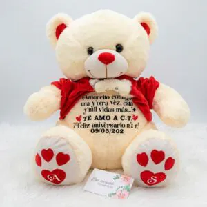 Oso de peluche beige con polo rojo levantado, mostrando un largo mensaje de aniversario personalizado en el vientre y huellas de corazón con iniciales.