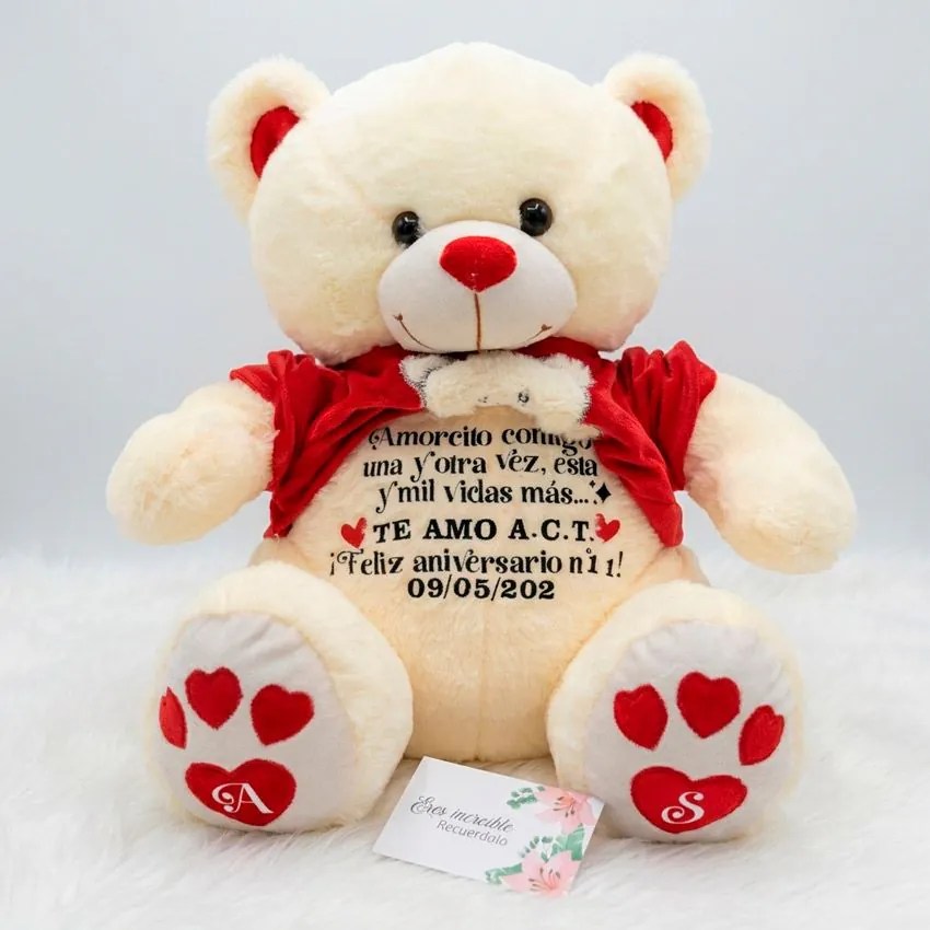 Oso de peluche beige con polo rojo levantado, mostrando un largo mensaje de aniversario personalizado en el vientre y huellas de corazón con iniciales.