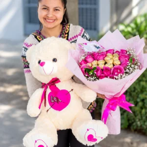 Abrazo Inolvidable: Oso de peluche color crema con detalles fucsias y ramo de rosas rosadas con chocolates de envoltura dorada.