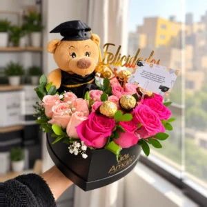 Osito graduado entre rosas rosadas y fucsias, chocolates Ferrero Rocher, follaje, tarjeta y letrero «Felicidades». Caja corazón Amalia.