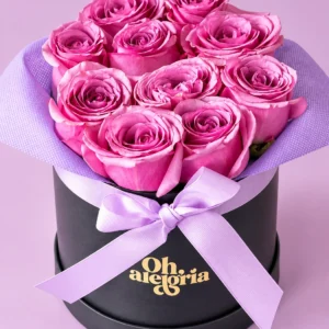 Cofre de Encanto: Rosas rosadas dispuestas en una caja redonda negra, con envoltura lavanda y un delicado lazo lila.