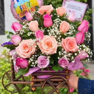 Jardín de Sueños: Rosas en tonos durazno, rosa y lila, flores blancas y follaje en una carretita decorativa.