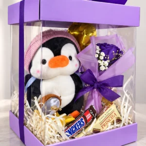 Pingüino Cómplice: Peluche de pingüino con gorrito, ramito con rosa morada, chocolates Snickers y Princesa, llavero y globo pequeño en caja transparente.