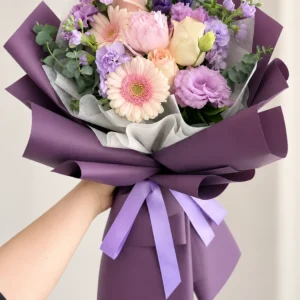 Poesía Pastel: Gerberas rosadas, rosas claras y flores lilas, violetas y blancas, con follaje.