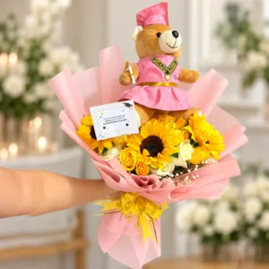Osita graduada con traje rosa, girasoles, rosas amarillas y blancas y flores de complemento. Incluye tarjeta y lazo dorado.
