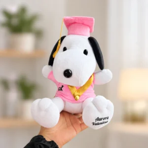 Snoopy con birrete y traje rosa, detalles dorados y bolsa de regalo incluida.